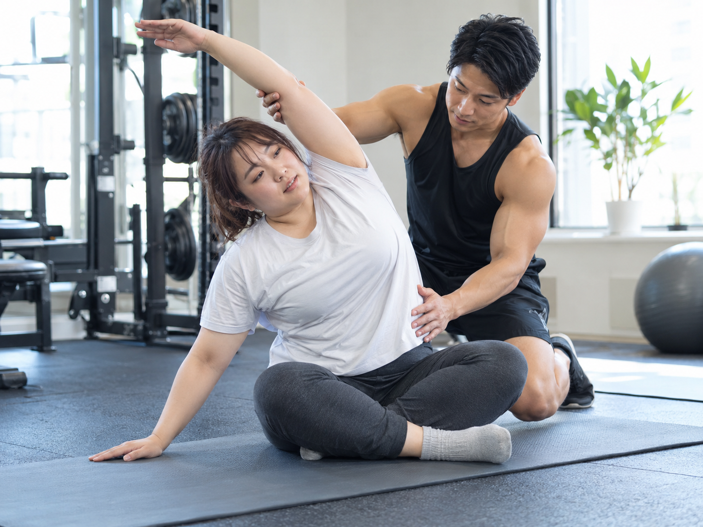 トレーナーのサポートを受けながら座って体を横に伸ばす女性のイメージ